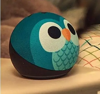 El mismo parlante esférico con una funda estampada de búho en tonos turquesa y blanco, apoyado sobre un velador